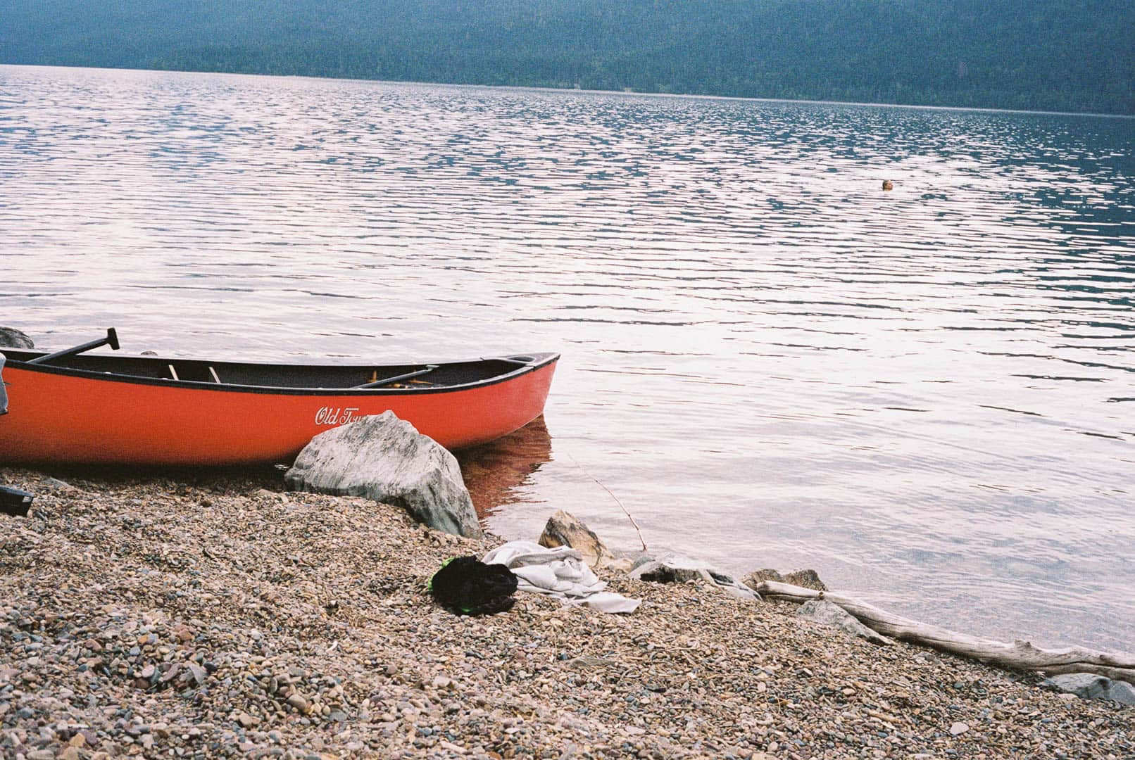 A bright red canoue on the lake shore with a pile of clothes nearby. In the distance you can see someones head sticking above the water