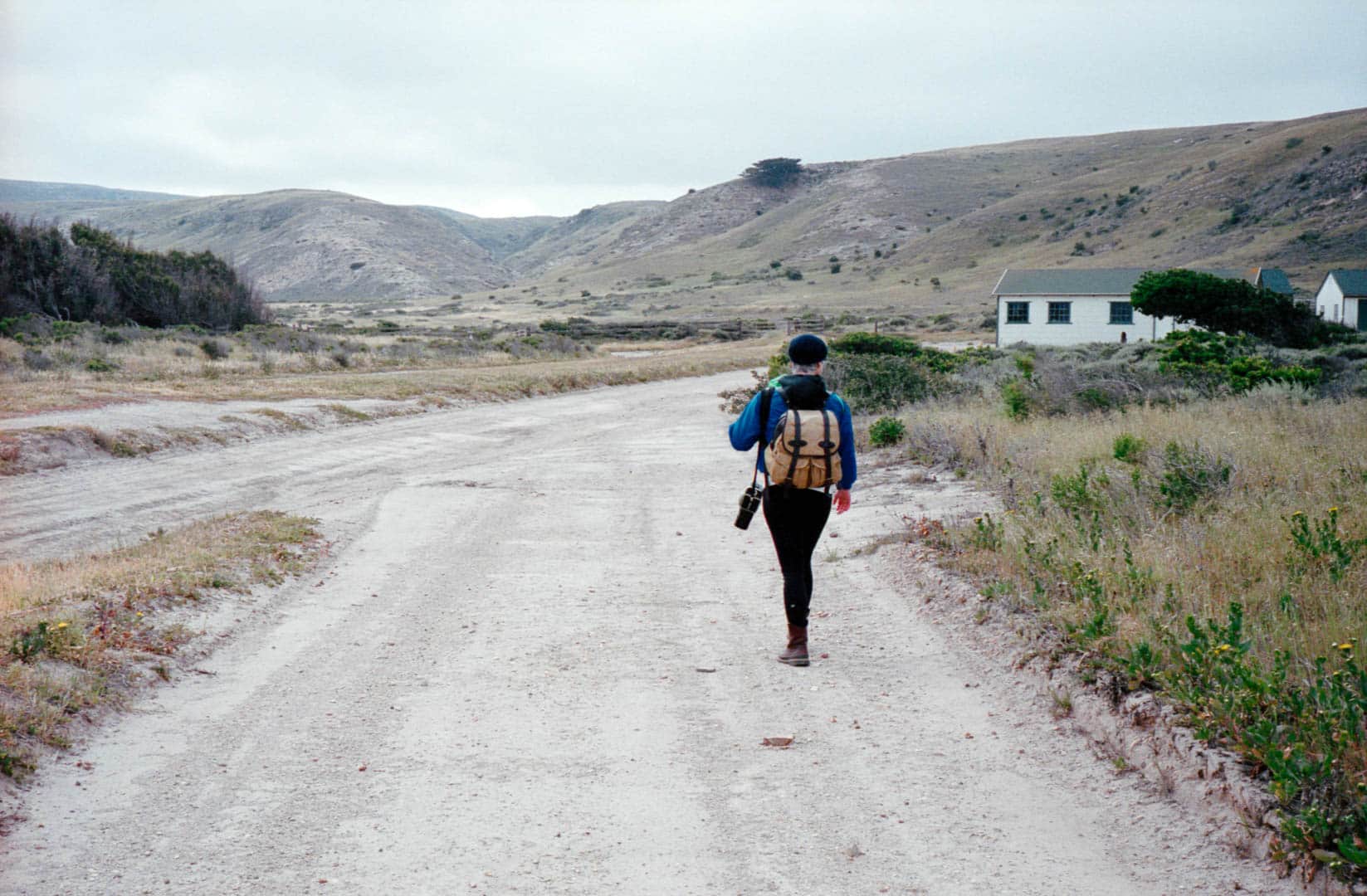 A woman walking into a landscape along a wide dirt path carrying a backpack and camera