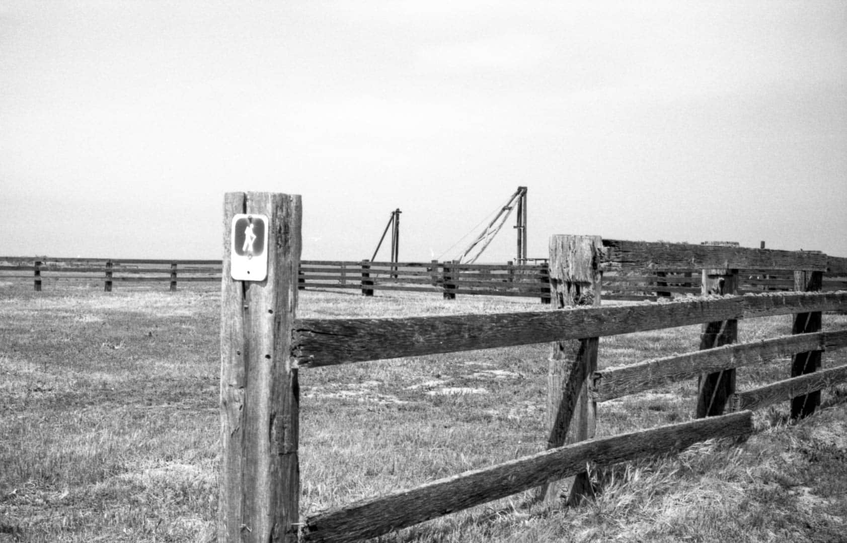 A hiking sign on a wooden fence along a grassy coastal trail with docks in the distance